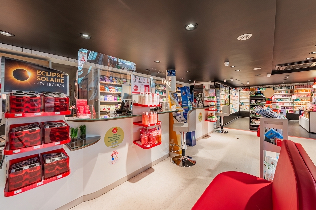 Une photo de l'intérieur de la pharmacie Sarrebourgeoise à Sarrebourg, France. L'image montre un comptoir de service incurvé avec un écran d'ordinateur et un terminal de paiement. Derrière le comptoir, une pharmacienne portant une blouse est partiellement visible. Les étagères au premier plan et en arrière-plan sont remplies d'une large gamme de produits de santé et de beauté de marques comme SVR et Caudalie. Un panneau d'affichage sur la gauche promeut les lunettes de protection solaire pour une éclipse. Un grand panneau suspendu fait de la publicité pour plusieurs marques. L'ambiance est lumineuse et moderne avec un plafond noir.
