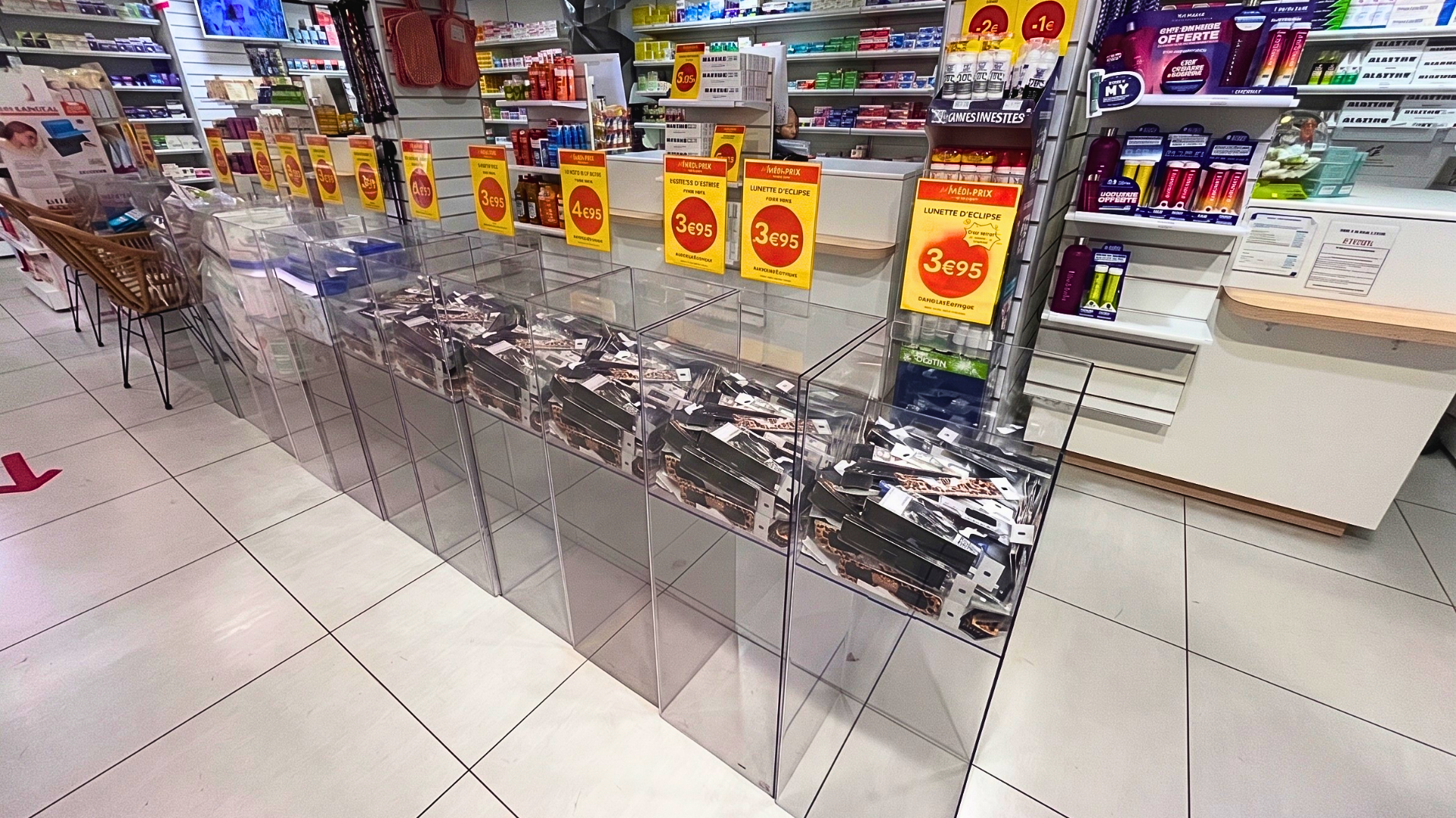 Une photo de l'intérieur d'une pharmacie moderne et lumineuse. L'allée principale est bordée de présentoirs en plexiglas transparent contenant des piles ordonnées de boîtes de lunettes d'éclipse solaire. Chaque bac est surmonté d'une étiquette jaune vif. À l'arrière-plan, on aperçoit le comptoir de service et des étagères remplies de médicaments. Le sol est carrelé avec des flèches directionnelles rouges.