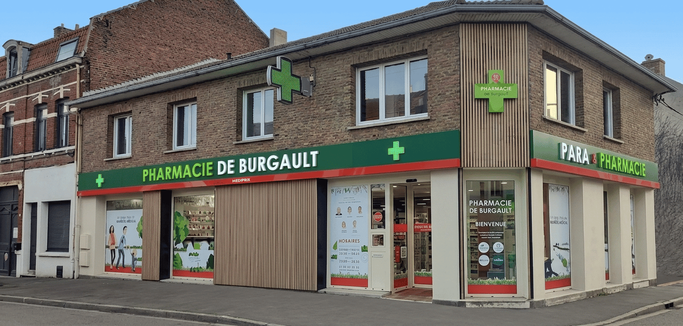 Façade d'angle de la Pharmacie de Burgault, associant briques anciennes et bardage en bois vertical clair. Les enseignes lumineuses vertes affichent « PHARMACIE DE BURGAULT » et « PARA & PHARMACIE ». Les grandes vitrines au rez-de-chaussée sont habillées de vitrophanies illustrées et d'affiches d'horaires. Le trottoir au premier plan est entièrement dégagé et propre, débarrassé de tout mobilier urbain, poteaux ou panneaux de signalisation.